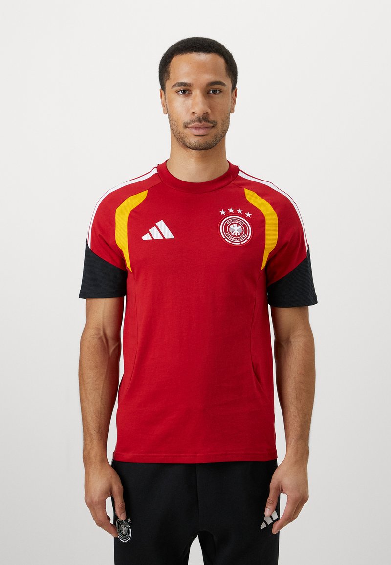 Νεαρός άνδρας που φο�ράει μια κόκκινη αθλητική φανέλα με κίτρινες και μαύρες λεπτομέρειες και μαύρες βερμούδες, στέκεται μπροστά σε έναν λευκό τοίχο.