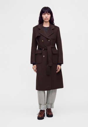 Femme portant un manteau croisé marron foncé avec ceinture, pantalon gris clair avec revers, et chaussures marron à lacets, debout devant un fond uni.