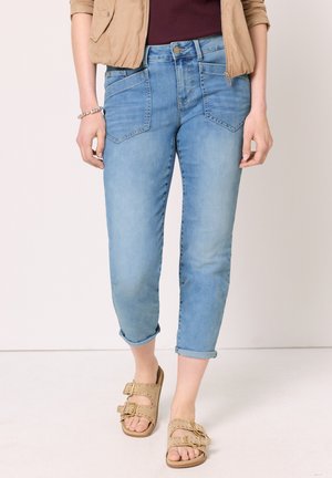 Kvinde iført lyseblå jeans med opsmøg, beige sandaler med dobbelt spænde, en vinrød top og en tan jakke med opkørte ærmer.