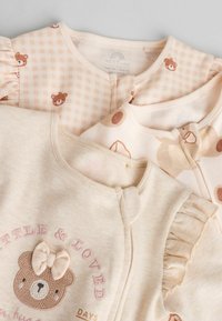 Tres prendas para bebés en suaves tonos beige: una presenta impresiones de ositos, otra tiene un patrón de cuadros, y la última tiene mangas con volantes y acentos bordados.