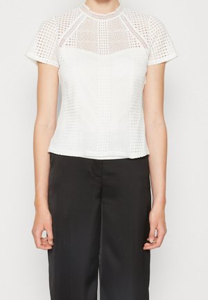 Kvinde iført en hvid kortærmet bluse i blonder med høj hals og sorte satin bukser med vide ben.