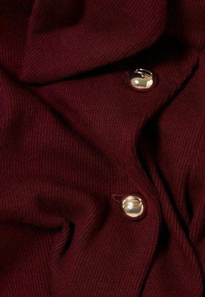 Tessuto a coste bordeaux con una morbida consistenza, caratterizzato da bottoni rotondi dorati. Il materiale ha un drappeggio liscio e flessibile con cuciture visibili.