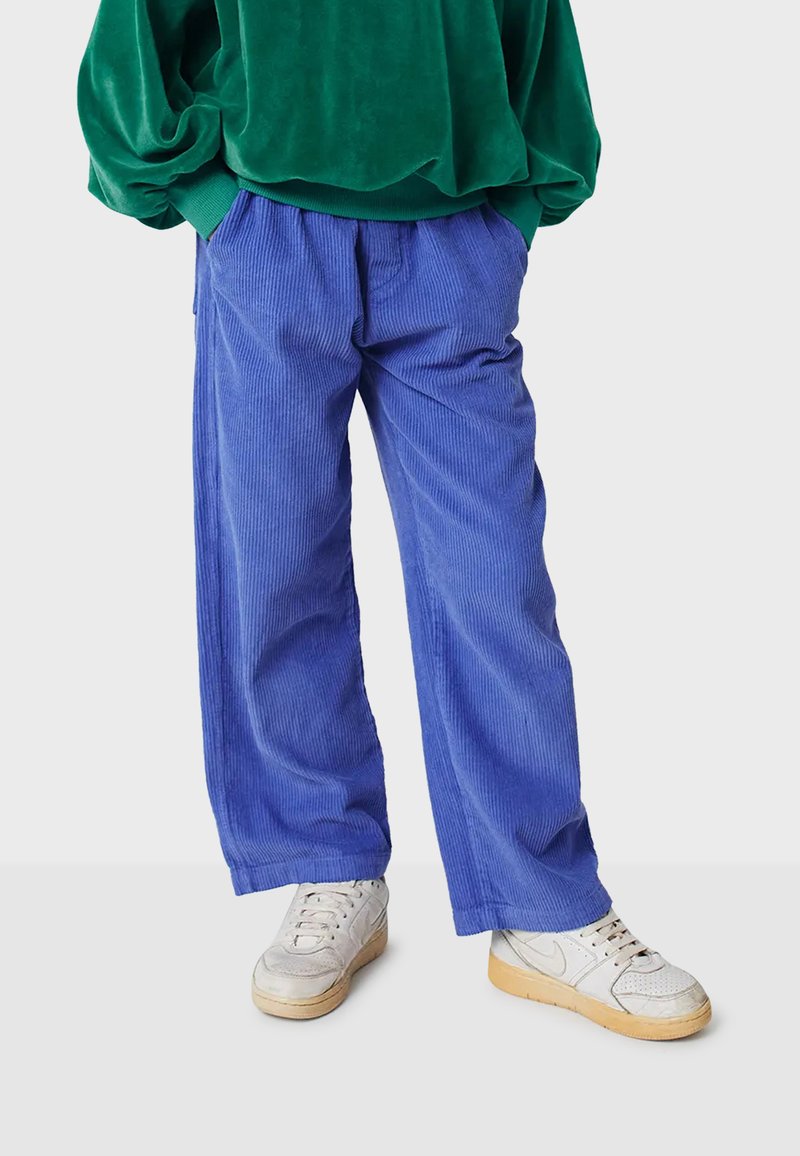 Blauwe corduroy broek met een relaxte pasvorm en verticale ribbels, gecombineerd met witte en grijze sneakers. Het bovenstuk is een groene oversized sweatshirt.