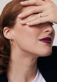 Złote kolczyki w kształcie obręczy z gładkim wykończeniem noszone są na lewym uchu. Ręka częściowo zakrywa twarz, podkreślając pierścionek z drobnymi akcentami.