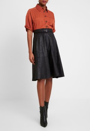 Kobieta ubrana w rdzawobrązową koszulę na krótkie rękawy z guziczkami, czarną spódnicę A-liniową o wysokim staninie z teksturą oraz pasek, a także czarne botki za kostkę.