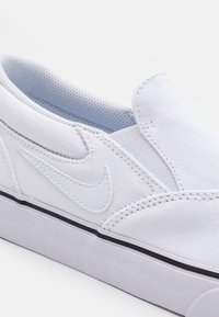 Baltie kanvas slip-on sporta apavi ar teksturētu virsmu, ar izteiktu baltu Nike logo un melnu gumijas zoli.
