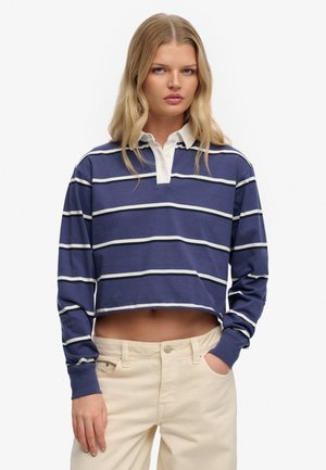 Ung kvinde iført en blå- og hvidstribet afkortet rugbytrøje og beige bukser, stående med den ene hånd i lommen mod en hvid baggrund.