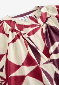 Blouse bordeau et crème avec un motif géométrique, des détails plissés le long du col, et une texture de tissu douce. Étiquette de marque visible.