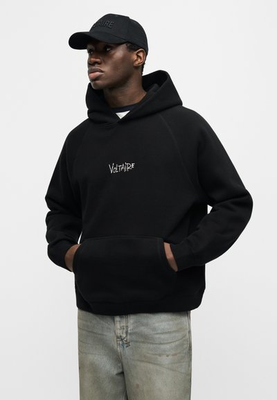 Νεαρός άνδρας που φοράει μαύρη φούτερ με κουκούλα με το κείμενο «Voltaire» και μαύρο καπέλο, στέκεται με τα χέρια στις τσέπες του φούτερ, με ανοιχτόχρωμο τζιν.