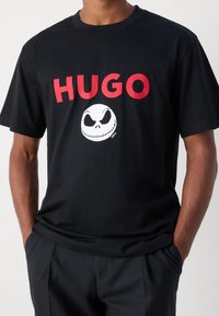 Musta puuvillapaita, jossa on rohkea punainen "HUGO" -teksti ja valkoinen Jack Skellingtonin kasvokuva keskellä, lyhyet hihat, pyöreä kaula-aukko.