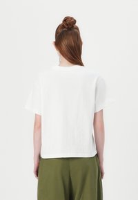 White cotton t-shirt with short sleeves, crew neck, relaxed fit, and a straight hem, viewed from the back. Paired with olive green pants.