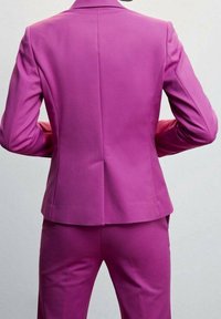 Personne portant un costume tailleur magenta vif vue de dos, les mains posées sur les hanches devant un fond uni.