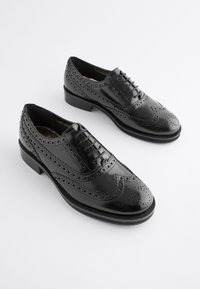 Scarpe brogue in pelle nera con perforazioni dettagliate, tacco basso e finitura liscia e lucida. Include lacci e design classico a punta alata.