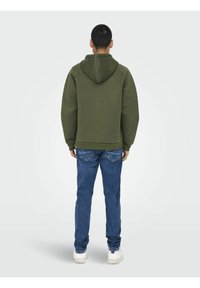 Olivgrön huvtröja med raglanärmar, tillsammans med blå denimjeans. Huvtröjan har en känguruficka och ribbad fåll.