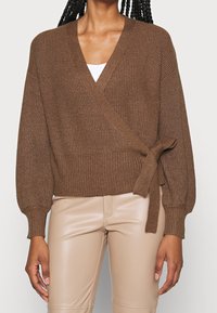 Pull cache-cœur côtelé marron en matière douce, avec un décolleté en V profond, des manches longues et un détail de taille nouée.