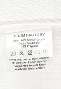 Vit tygetik med texten "DENIM FACTORY". Innehåller skötselråd och materialkomposition: 60% bomull, 40% polyester.