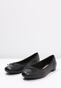 Ballerines en cuir noir avec des orteils ronds, présentant un petit nœud sur le devant et une semelle fine et plate. Texture lisse et design classique.