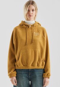 Blondynka w musztardowej polarowej bluzie z kieszenią z przodu i białym haftowanym napisem, w połączeniu z niebieskimi dżinsami, na jednolitym tle.