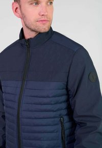 Marineblauwe jas met een gladde bovenkant en een gevoerde onderkant, voorzien van een rits aan de voorkant en een logo op de linkermouw.