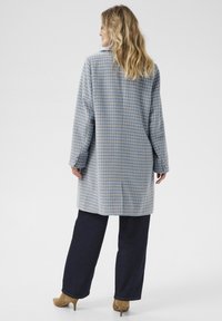 Cappotto lungo e oversize in un motivo a quadri azzurri chiari con accenti beige, tessuto testurizzato e tasche laterali. Abbinato a pantaloni in denim scuro.
