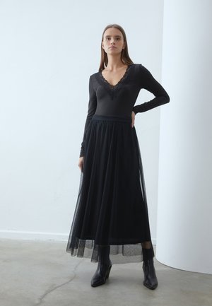 Bodysuit noir à manches longues avec un col en dentelle, associé à une jupe maxi en tulle noire et des bottines à talons noires, sur fond de mur clair.