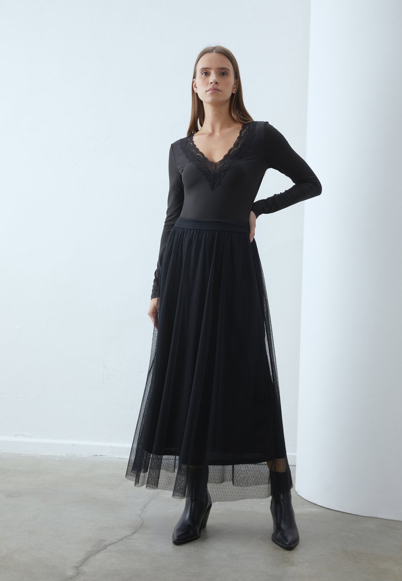 Sort langærmet bodysuit med blondeudskæring, sammen med en sort tulle maxi-skirt og sorte hælede ankelstøvler, sat imod en lys væg.