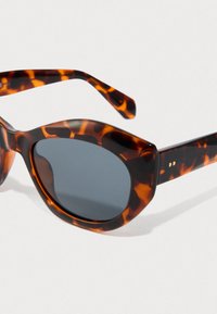 Occhiali da sole con motivo tartarugato, lenti ovali scure e montatura spessa, posati su una superficie bianca.