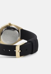 Black rubber watch strap with a gold buckle, paired with a gold-tone case. The back features a clear, metallic surface.