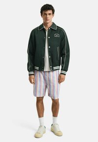 Grüne leichte Bomberjacke mit weißen Paspeln, gestreifte Shorts in Pastellfarben, weiße Sneaker und gerippte Socken. Lässiges Outfit.