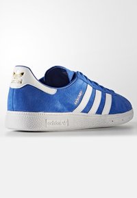 Blå mockalöparsko med tre vita ränder och vit gummisul. Har guldfärgade bokstäver och en texturerad tåkappa.