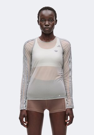 Femme portant un haut à manches longues translucide sur un soutien-gorge de sport blanc et un short de sport marron avec un fond neutre, regardant devant avec une expression neutre.
