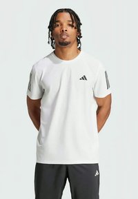 T-shirt de sport à manches courtes blanche avec le logo Adidas noir et trois bandes noires sur l'épaule. Le matériau semble léger avec une texture lisse.