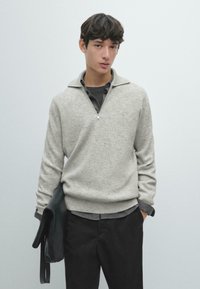 Hellgrauer Strickpullover mit Reißverschluss am Kragen, �übereinander getragen mit einem dunkelgrauen Hemd, kombiniert mit schwarzen Hosen. Glatte Textur, lässige Passform.