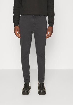 Mann trägt dunkelgraue schmal geschnittene Hose, schwarzen Pullover, weiße Socken und schwarze Ledermokassins, steht vor einem schlichten hellen Hintergrund.