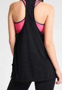 Svart atletisk linne med racerback-design, som har en lös passform och sidoklipp. Underneath syns en rosa sportbehå.