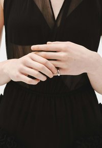 Vestido negro transparente con una cintura ajustada, que presenta un acento con volantes. Las manos lucen un anillo plateado simple de forma ovalada. Textura de piel suave.