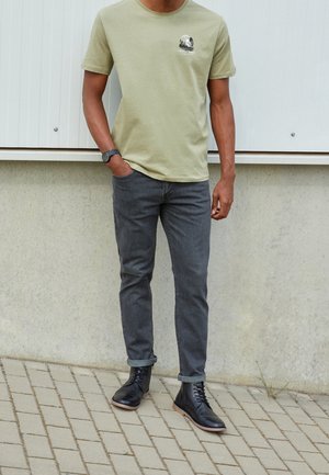 Mand iført lysegrøn t-shirt med grafik, mørkegrå opkrogede jeans, sorte læderstøvler og sort armbåndsur, stående udendørs.