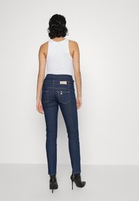 Jeans azul oscuro con un diseño ajustado, bolsillos traseros y una cinturilla texturizada. Llevados con una blusa blanca sin mangas y botas de tacón negro.