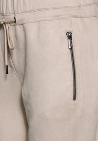 Beige joggers gemaakt van zachte stof, met een elastische tailleband, trekkoord en een zijzak met rits en zilverkleurige hardware.