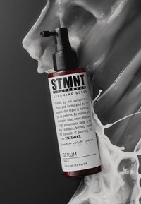 Röd flaska med STMT grooming-serum och svart pump. Etiketten har fetstilta texter, detaljerade beskrivningar och signaturer. Omgiven av vit vätska.