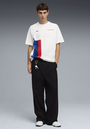Młody mężczyzna w białej koszulce Puma BMW Motorsport z niebieskimi i czerwonymi paskami, czarnych szerokich spodniach, białych trampkach oraz z kluczami przypiętymi do spodni.