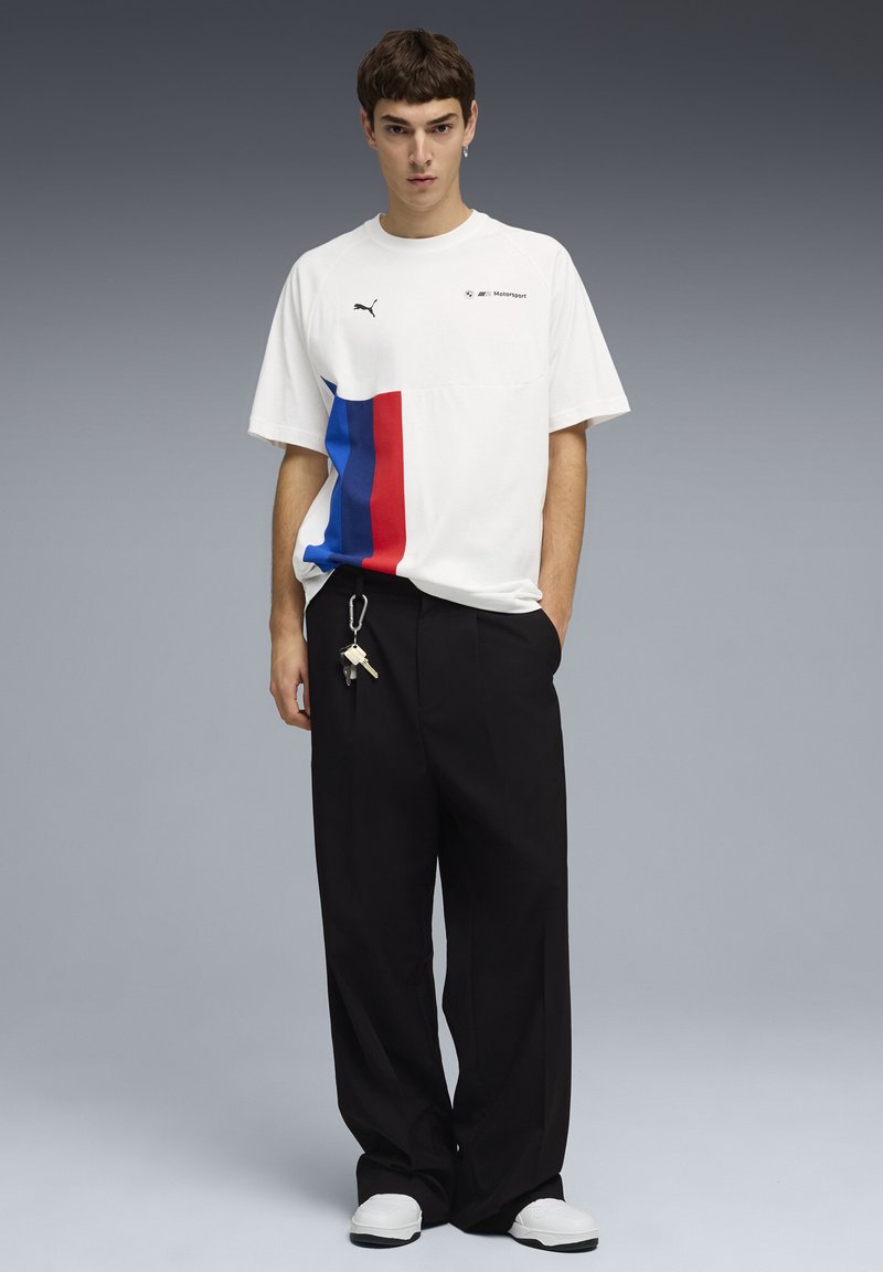 Νεαρός άνδρας με λευκό t-shirt Puma BMW Motorsport με μπλε και κόκκινες ρίγες, μαύρο φαρδύ παντελόνι, λευκά αθλητικά παπούτσια και κλειδιά κρεμασμένα στο παντελόνι.