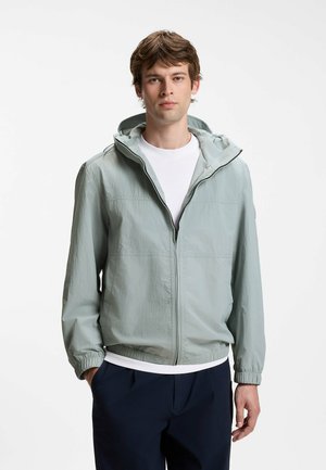 Young man wearing light gray hooded zip-up jacket over a white shirt and dark pants, standing with hands in pockets against a plain background.