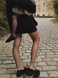 Sorte fiskenet strømpebukser der komplementerer en kort sort nederdel, parret med klumpede sorte sko, mod en brostensbelagt sti med grønne planter.