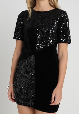 Femme portant une robe courte noire avec un design en patchwork moitié velours, moitié paillettes, manches courtes.