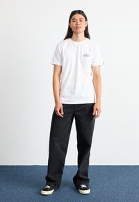 Weißes Baumwoll-T-Shirt mit einem kleinen Grafik auf der linken Brust, kombiniert mit schwarzen weit geschnittenen Jeans und schwarzen Sneakers mit cremefarbenen Akzenten.