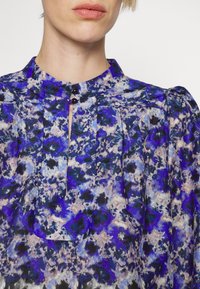 Gros plan d'une personne portant un chemisier à motifs floraux violet, bleu et noir avec un petit col et deux boutons noirs près du cou.