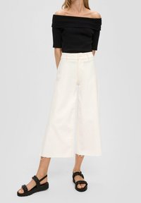 Donna con top nero off-shoulder, pantaloni bianchi a vita alta e gamba ampia, e sandali con plataforma intrecciati neri, in piedi su uno sfondo grigio.