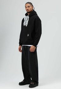 Zwarte hoodie met een bandana-print accent op de borst, gecombineerd met loszittende zwarte jeans en zwarte schoenen met veters detail.