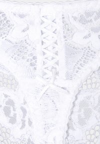 Lingerie en dentelle blanche avec des motifs floraux complexes, un bord festonné et un détail central en dentelle style corset avec un petit nœud.